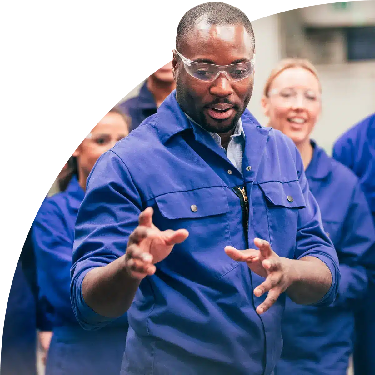 A man in blue coveralls and safety goggles speaks and gestures animatedly during a technical training session, with a group of similarly dressed colleagues watching attentively in the background.