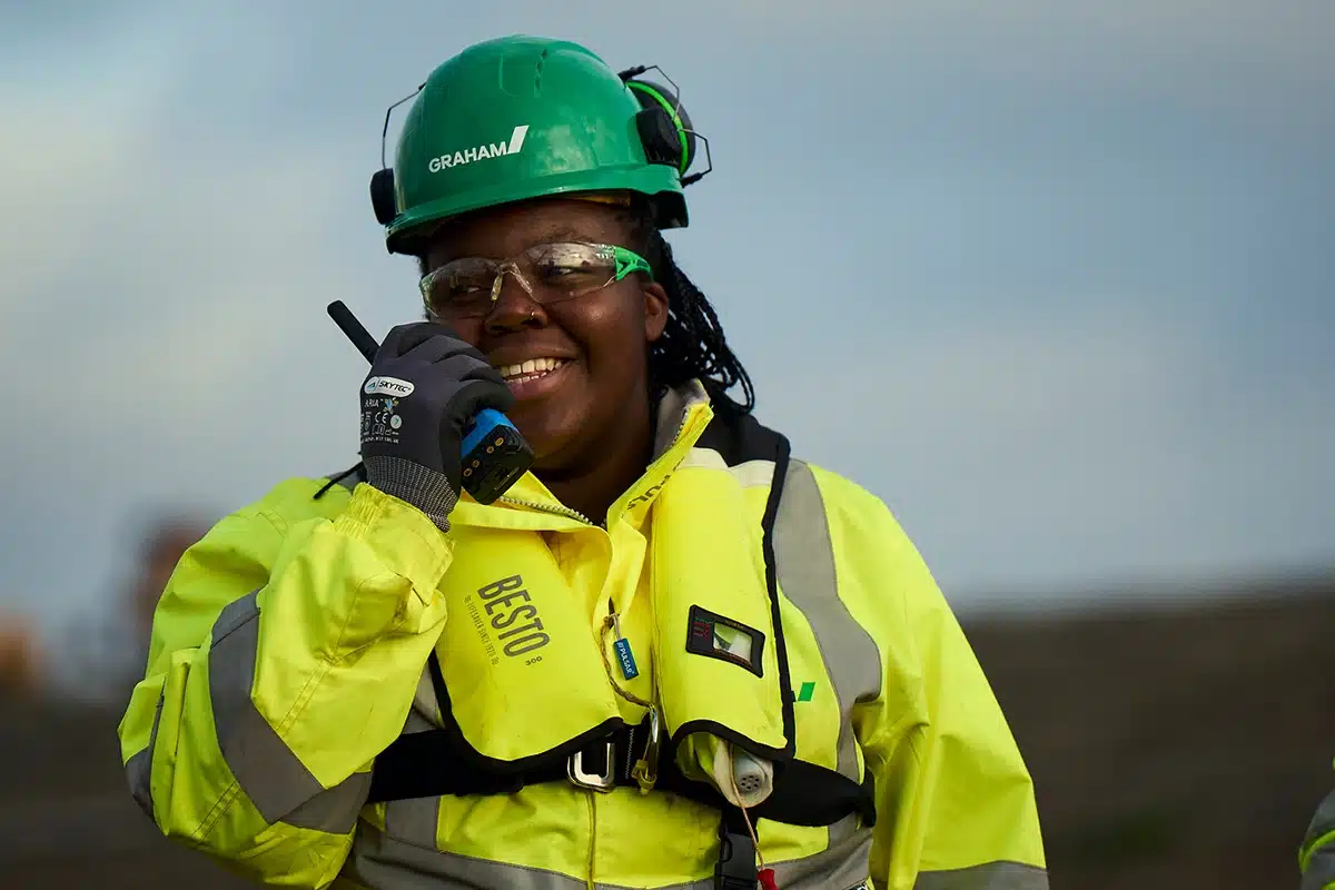 Smiling female offshore wind technician wearing green Graham safety helmet, safety glasses, and high-visibility yellow jacket with reflective strips, holding radio communication device against cloudy sky backdrop