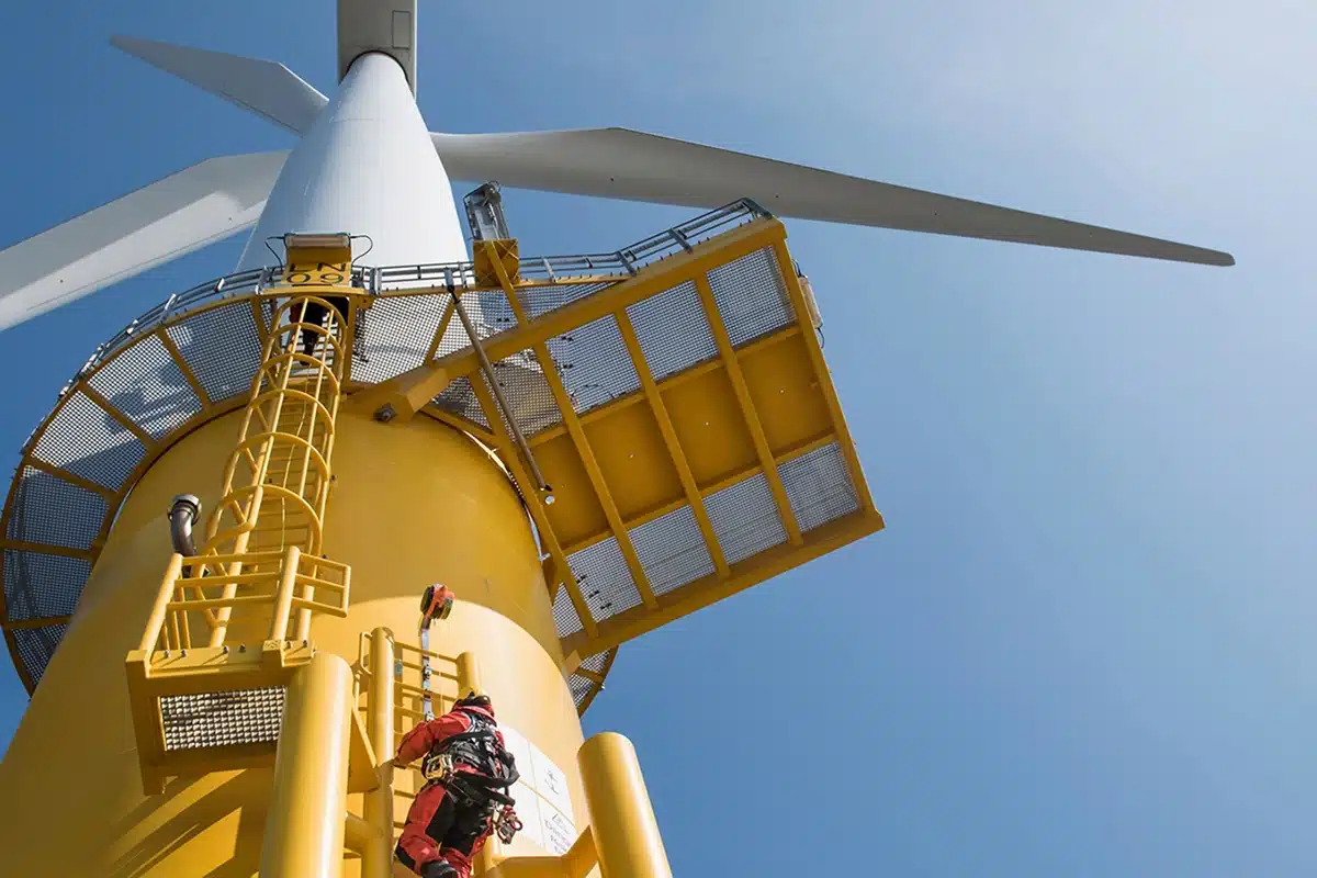 Technician in red safety gear climbing yellow maintenance ladder on offshore wind turbine tower, with white turbine blades visible against clear blue sky