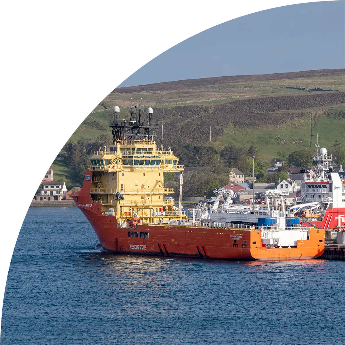 A red and orange offshore support vessel with yellow superstructure moored in a harbor, with a coastal town and rolling hills visible in the background. The image is presented in an oval frame.