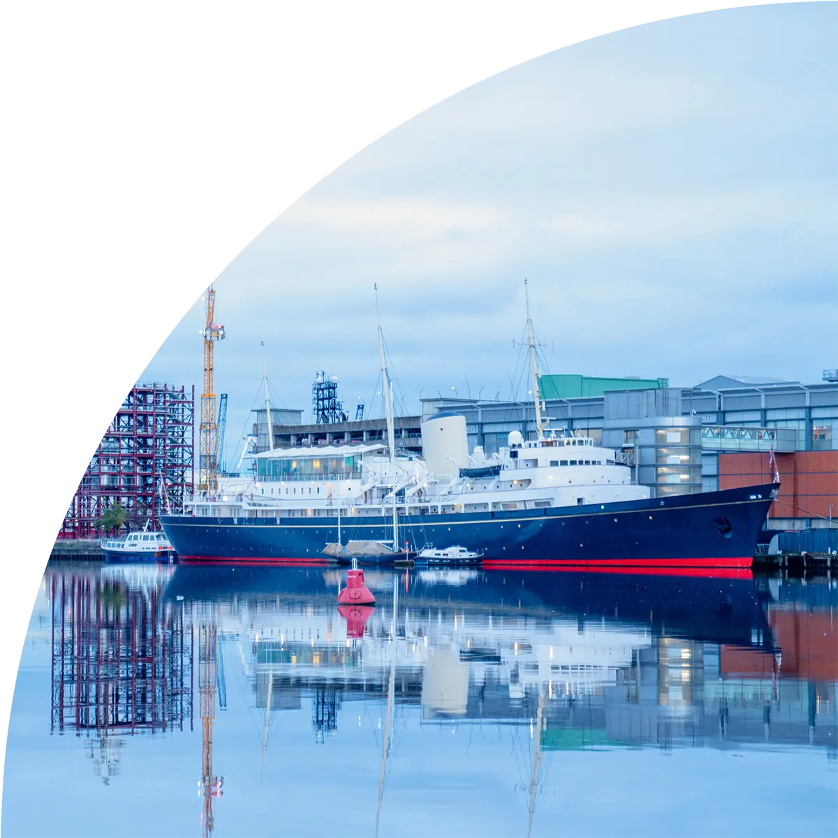 A large white and blue passenger ship moored at a harbor dock with perfect reflections in calm water. The vessel has a red hull stripe, and industrial port structures including cranes and warehouses are visible in the background.