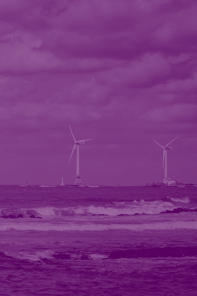 Two white offshore wind turbines standing in ocean water beyond rocky shoreline with waves breaking on dark rocks, under partly cloudy blue sky with construction or service vessels visible near the turbines
